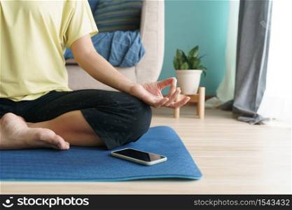 Hand of woman in lotus pose while sitting at mat yoga, Asian women practice yoga. With an online Trainer on mobile phone at home. Concept of relaxation and meditation