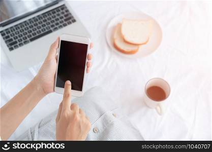 Hand of working woman holding and touching the black smartphone screen. Asian woman sitting on a white cloth while having bread, coffee and laptop.
