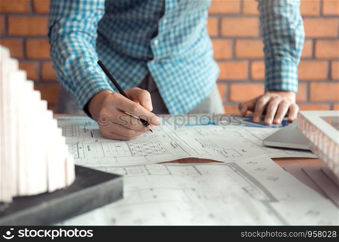 Hand of young engineering man drawing on blueprint with model on desk.