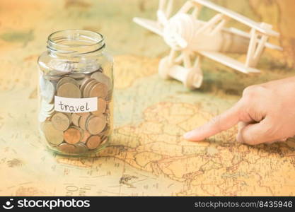 Hand pointing attractions in a map on a table with a glass jar with coins and travel inscriptions and a wooden toy plane nearby.