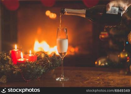 Hand pouring champagne in glass. Christmas tree and burning fireplace on the background