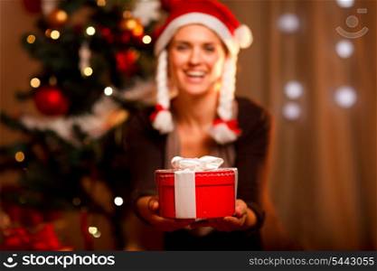 Hand presenting gift box and smiling woman and Christmas tree in background&#xA;