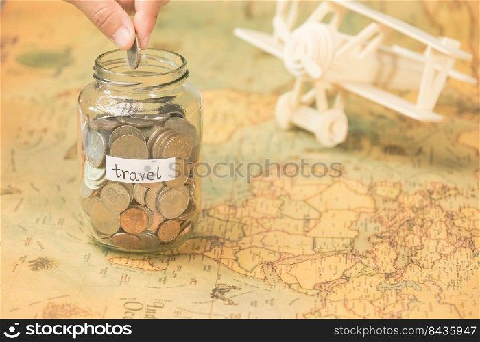 Hand putting coins in a clear jar on the table to save for travel. Glass jar with coins and an inscription travel on the world map with wooden toy plane on the table.