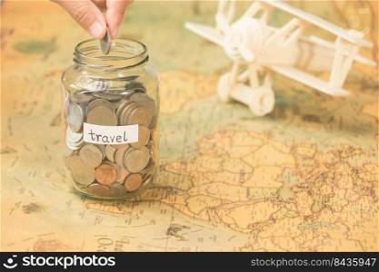 Hand putting coins in a clear jar on the table to save for travel. Glass jar with coins and an inscription travel on the world map with wooden toy plane on the table.