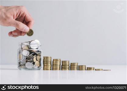 hand putting money in glass jar near decreasing stacked coins