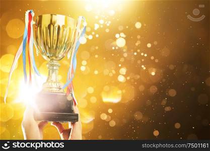 Hand raised, holding gold cup with abstract shiny lights . award and victory concept .