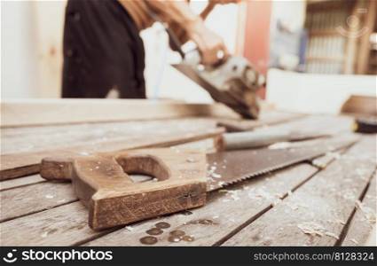 Hand saw with a wooden handle on blur carpenter working with electric wood planer. Carpenter tools. Hand saw and sawdust on wood table at outdoor workshop. Craftsman making woodwork. Handwork concept.