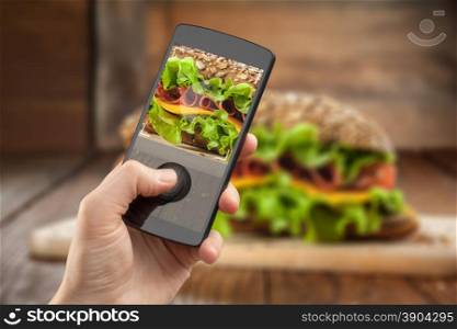 Hand taking photo of sandwich with smatphone on the wooden table with slices of tomatoes, ham, cheese and lettuce