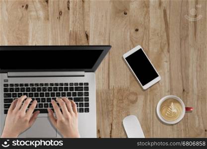 hand typing on laptop, smartphone, mouse, coffee cup on wooden desk - top view