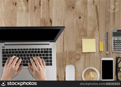 hand typing on laptop, smartphone, mouse, coffee cup on wooden desk - top view