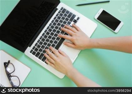 Hand using laptop on creative flat lay workspace desk with smartphone and stationery on green background