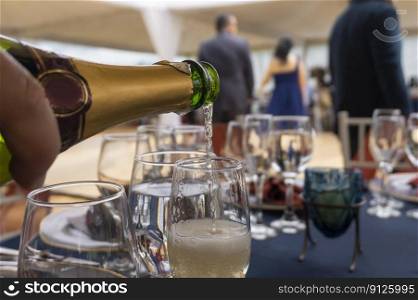 Hand with bottle of ch&agne filling a crystal glass on a table with blue tablecloth with background of people dancing at a party. Hand with bottle of ch&agne pouring a crystal glass on a table with a blue tablecloth with a background of people dancing