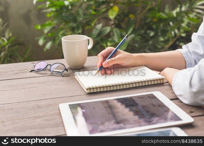 Hand woman writing notebook on wood table with cup coffe and tablet.