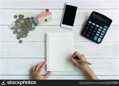 Hand writing on notebook, house model, coins and calculator on white wooden background, savings plans for housing financial concept, copy space, top view