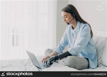 Handicapped freelancer has online meeting sitting on her bed. Young caucasian woman with artificial arm is working on laptop in bedroom. Girl is using headphones and microphone and chatting.. Handicapped freelancer has online meeting sitting on her bed. Girl using headphones and microphone.