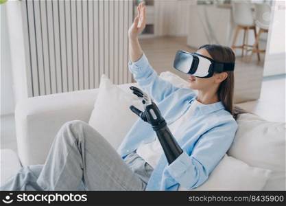 Handicapped girl in virtual reality glasses at home. Disabled person rehabilitation. European woman is getting used to modern bionic prosthesis using vr headset. Electronic technology in medicine.. European woman is getting used to modern bionic prosthesis using vr headset. Electronic technology.