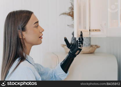 Handicapped girl is opening cupboard with electronic cyber hand. Young european woman with artificial arm at home. Functions of modern bionic prosthesis. Routine of disabled person concept.. Handicapped girl is opening cupboard with electronic cyber hand. Routine of disabled person.