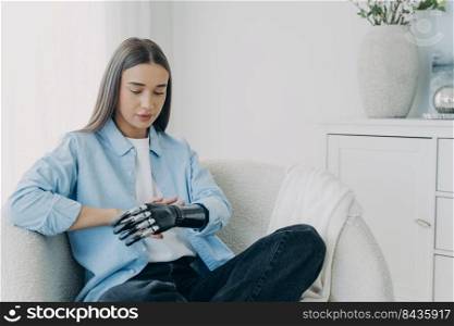 Handicapped girl looks and touches her bionic arm. Young pensive european woman with cyber hand at home. Modern bionic prosthesis. Futuristic technology of artificial limbs.. Handicapped european girl looks and touches her bionic arm. Modern bionic prosthesis.
