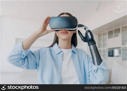 Handicapped girl with cyber arm in vr glasses at home. Disabled person gets rehabilitation. Young european woman sitting in living room on couch is excited. Robotic technologies and bionic prosthesis.. Handicapped girl with cyber arm in vr glasses at home. Disabled person gets rehabilitation.