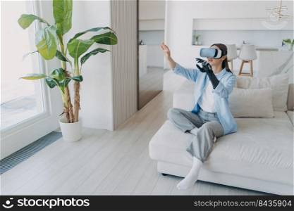 Handicapped girl with cyber arm in vr glasses at home. Technology for recovery. Disabled person gets rehabilitation. Young european woman sitting in living room is excited. Lady touches the vision.. Handicapped girl with cyber arm in vr glasses at home. Technology for recovery and rehabilitation.