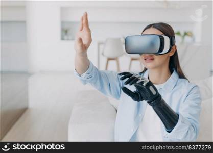 Handicapped girl with cyber bionic hand in virtual reality glasses at home. Disabled person gets rehabilitation. Young european woman sitting in living room on couch. Excited woman touches the vision.. Handicapped girl with cyber bionic hand in virtual reality glasses gets rehabilitation.