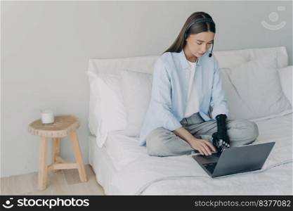 Handicapped remote worker. Concentrated girl has online meeting sitting on her bed at home. Young caucasian woman with artificial arm is working on laptop in bedroom. Manager or virtual assistant.. Handicapped remote manager or assistant. Concentrated girl has online meeting at home.