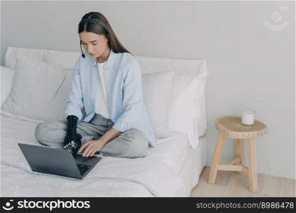 Handicapped remote worker. Concentrated girl has online meeting sitting on her bed at home. Young caucasian woman with artificial arm is working on laptop in bedroom. Manager or virtual assistant.. Handicapped remote manager or assistant. Concentrated girl has online meeting at home.