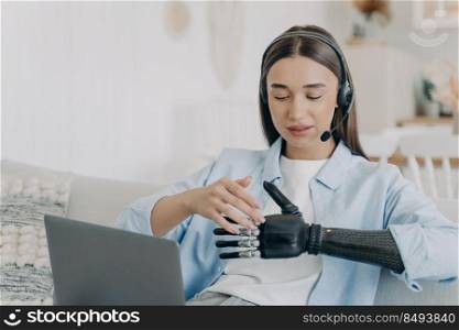 Handicapped young woman with artificial arm in headset and microphone at home. Freelancer has online meeting on pc. Happy european handicapped girl has electronic prosthesis. Remote work at home.. Handicapped young woman with artificial arm in headset at home. Freelancer has online meeting.