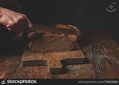 Hands cutting rye bread on kitchen wood plate with a chef holding gold knife for cut.. Hands cutting rye bread