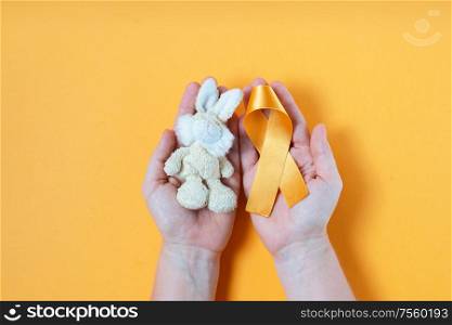 Hands holding children&rsquo;s toy rabbit with a Childhood Cancer Awareness yellow ribbon on yellow background with copy space. Childhood Cancer Day February, 15. Childhood Cancer Awareness concept