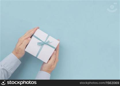 Hands holding Gift box with blue bow with copy space in craft paper over blue background. Top view, flat lay