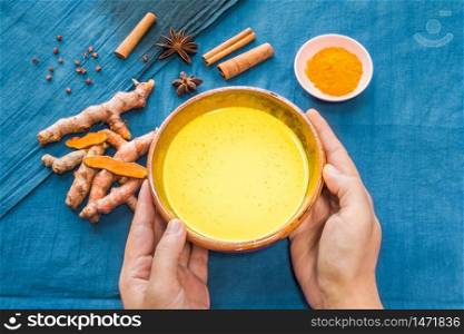Hands holding organic curcumin honey golden milk, Indian turmeric latte with ingredients on bright blue background. Top view exotic spices yellow natural drink healthy food concept