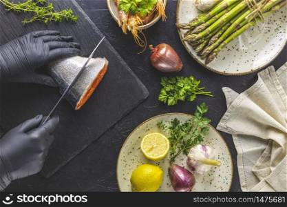 Hands in black gloves cut trout fish on black stone cutting board surrounded herbs, onion, garlic, asparagus, shrimp, prawn in ceramic plate. Black concrete table surface. Healthy seafood background.