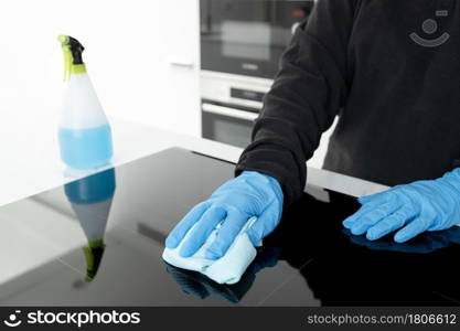 Hands in protective glove cleaning a modern electric stove with rag of a minimalist kitchen