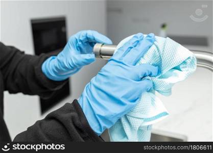 Hands in protective glove cleaning a modern tap with rag of a minimalist domestic kitchen