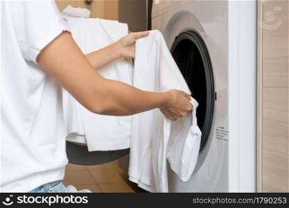 Hands of Asian woman loading white color clothes into washing machine in kitchen at home. Close up. Laundry concept.