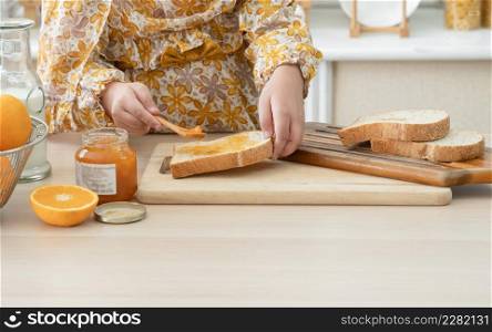 Hands of Muslim kid girl wear hijab holding spoon and spreading jam on sliced whole wheat bread for preparing breakfast on kitchen table at home with oranges and milk