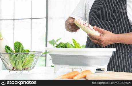 Hands of senior man washing and rubbing corn at home. Vegetables soak with water in basin. White radish, green vegetables, tomato, carrots in basket. Healthy food and cleaning before cooking