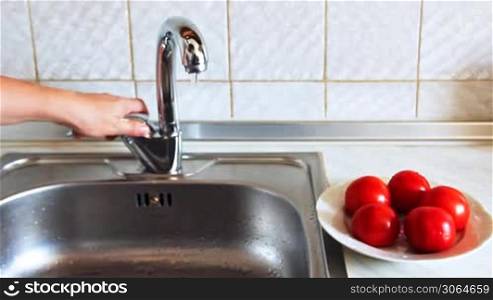 hands of woman open faucet with cold water and thoroughly wash red tomato, then put it in plate