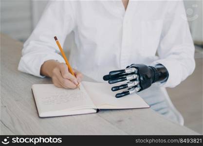 Hands of woman which is taking notes with pencil. Cyber arm and healthy limb on the notebook. Handicapped young woman is getting remote education. Disabled european girl is writing at the desk.. Hands of woman which is taking notes with pencil. Cyber arm and healthy limb on the notebook.