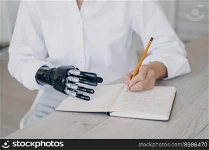 Hands of woman which is taking notes with pencil. Cyber arm and healthy limb on the notebook. Handicapped young woman is getting remote education. Disabled european girl is writing at the desk.. Hands of woman which is taking notes with pencil. Cyber arm and healthy limb on the notebook.