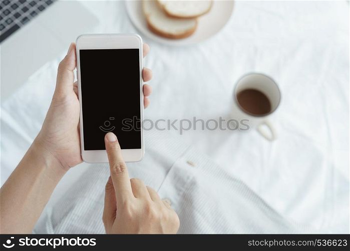 Hands of working woman holding and using smart phone on desk background. On the desk have cup coffee, breads and laptop.