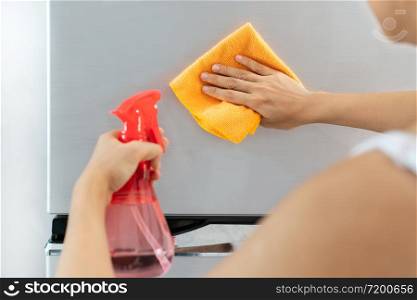 Hands of young Asian woman cleaning stainless steel refrigerator with cloth and spray bottle at home