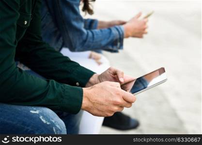 Hands of young unrecognizable people using smartphone and tablet computers outdoors in urban background. Women and men sitting on a bench in the street wearing casual clothes.