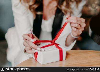 Hands of young woman with tender manicure wrapped gift box, wants to see what present she recieved. Unrecognizable female ties on bow at wooden table. Holiday decoration. Parcel in girls hands