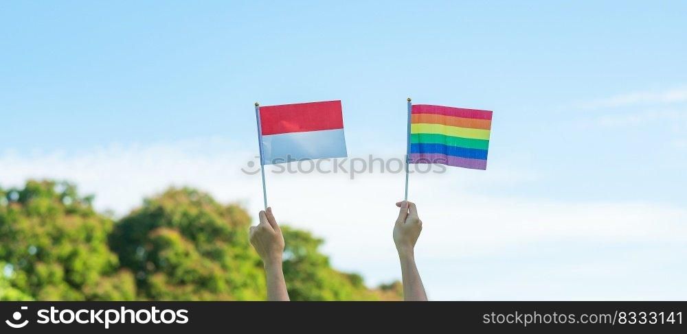 hands showing LGBTQ Rainbow and Indo≠sia flag on nature background. Support Lesbian, Gay, Bisexual, Trans≥nder and Queer comμnity and Pride month concept