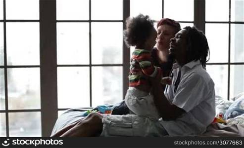 Handsome african american dad with dreadlocks lifting curly mixed race toddler son up and kissing his belly with love and tenderness while speding time together with caucasian mother at home. Interracial family lounging in the bedroom. Side view.