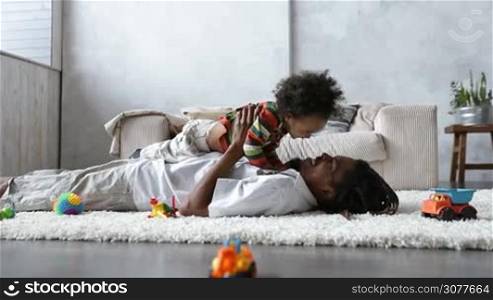 Handsome african american father with dreadlocks lying on his back on white woolen carpet and his curly mixed race toddler son lying on his chest. Side view of dad with young cute child spending time together at home over modern domestic interior.