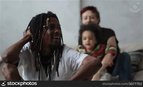 Handsome african american father with dreadlocks sitting on the floor in the living room and ready to hug his curly mixed race son over blurry laughing caucasian mother playing with child. Slow motion.