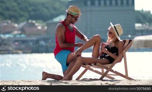 Handsome afro american man applying sunscreen on his girlfriend&acute;s leg on the beach. Beautiful young woman relaxing on sun lounger and drinking lemonade. Man applying skin care sun protect on woman tanned leg .